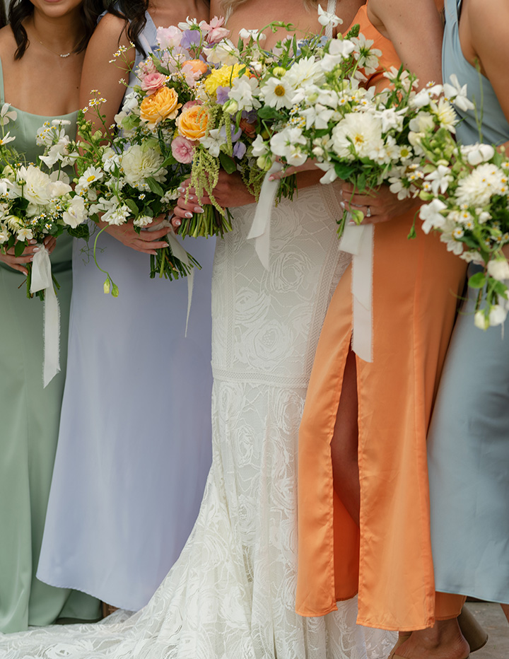 Bridesmaids in pastel dresses and a bride in a white gown hold vibrant bouquets with white, orange, and pink flowers. The mood is joyful and elegant.
