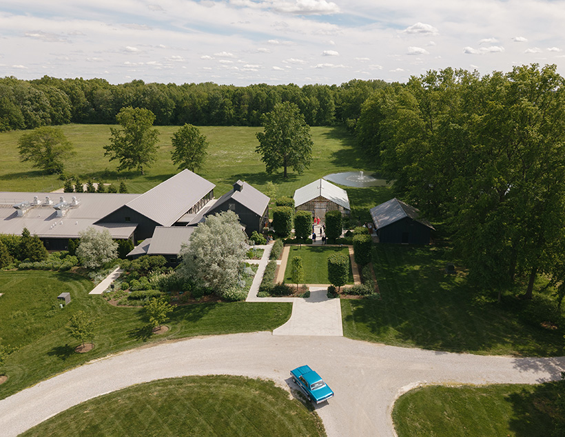 Aerial view of Jorgensen Farms Westerville Ohio with modern buildings, lush gardens, and a central fountain. A blue vehicle is parked on a gravel path under a clear sky.