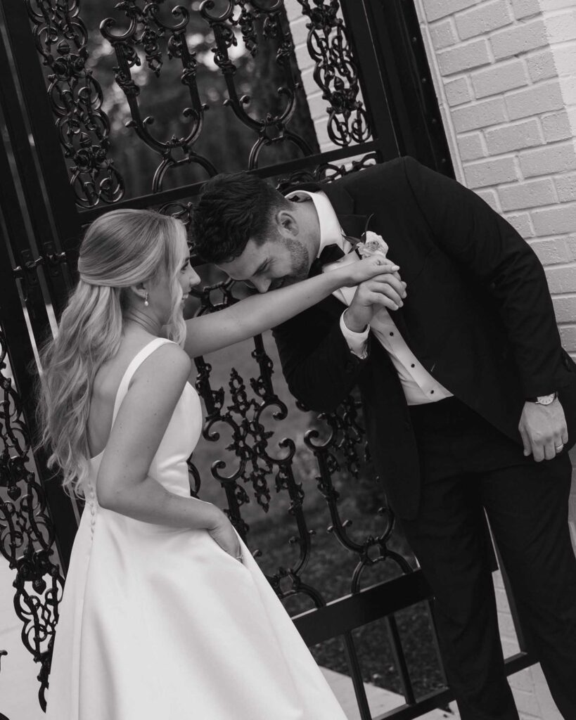 Franklin, Ohio couple shares an intimate moment in front of a decorative iron gate. The groom kisses the bride's hand tenderly. Both smile, exuding happiness.