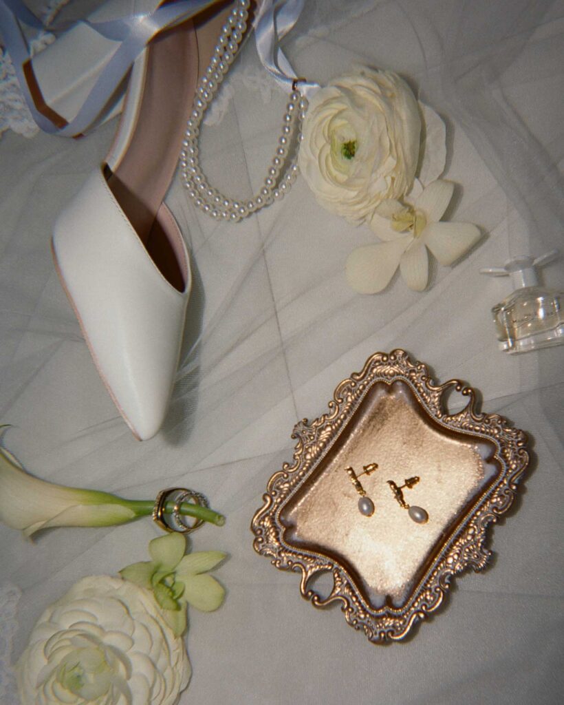 Elegant bridal flat lay with a white shoe, pearl necklace, flowers, gold-framed tray with earrings, and a perfume bottle on a soft fabric. Romantic mood.