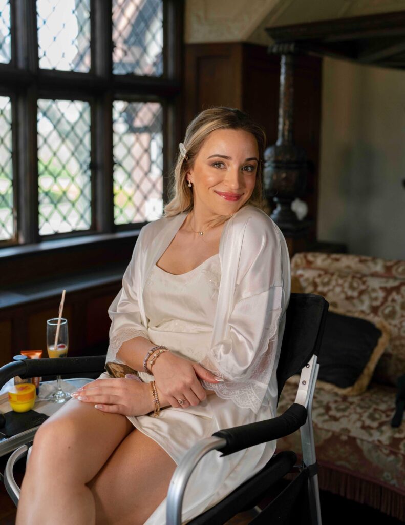 Smiling woman in a silky robe sits on a chair by a window with lattice panes, exuding warmth and relaxation. A drink and cozy decor enhance the serene scene.