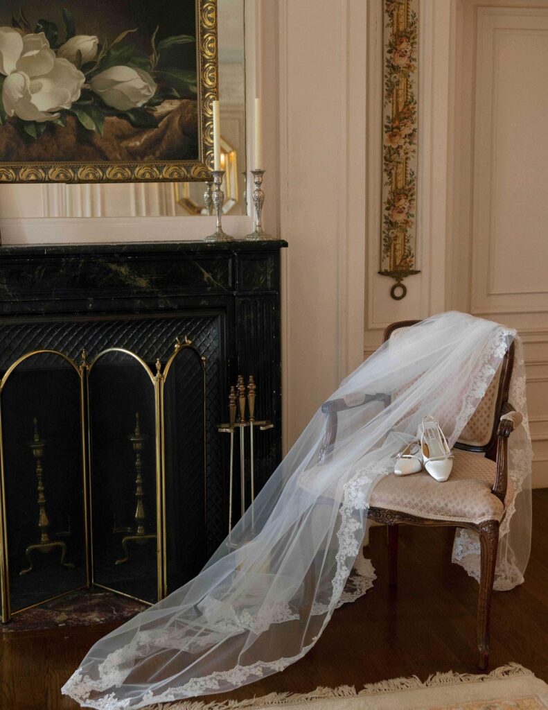 An elegant bridal veil and silver shoes on a vintage chair beside a dark, ornate fireplace. Floral artwork and soft lighting create a serene, romantic atmosphere.