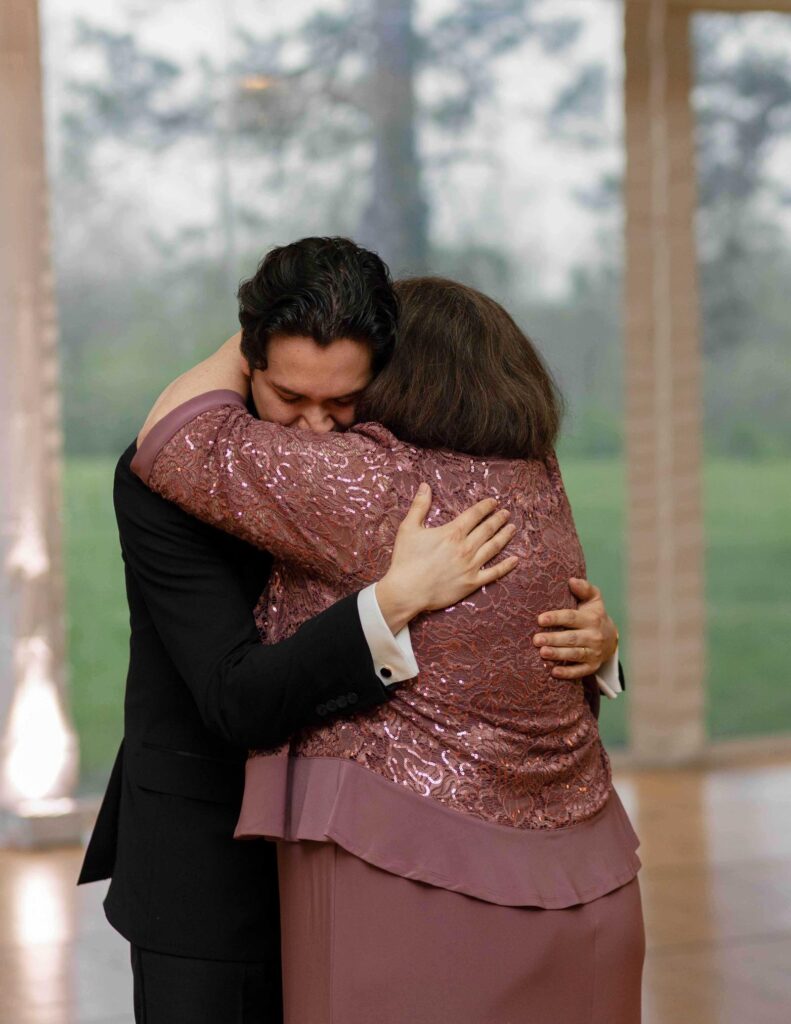A man in a black suit and a woman in a shimmering mauve dress share a heartfelt hug, conveying warmth and emotion in a softly lit room with large windows.