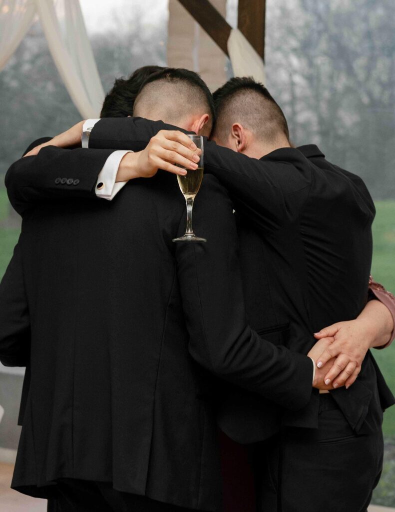 Three people in suits huddle closely, embracing each other warmly. One holds a champagne glass, conveying a celebratory, intimate moment.