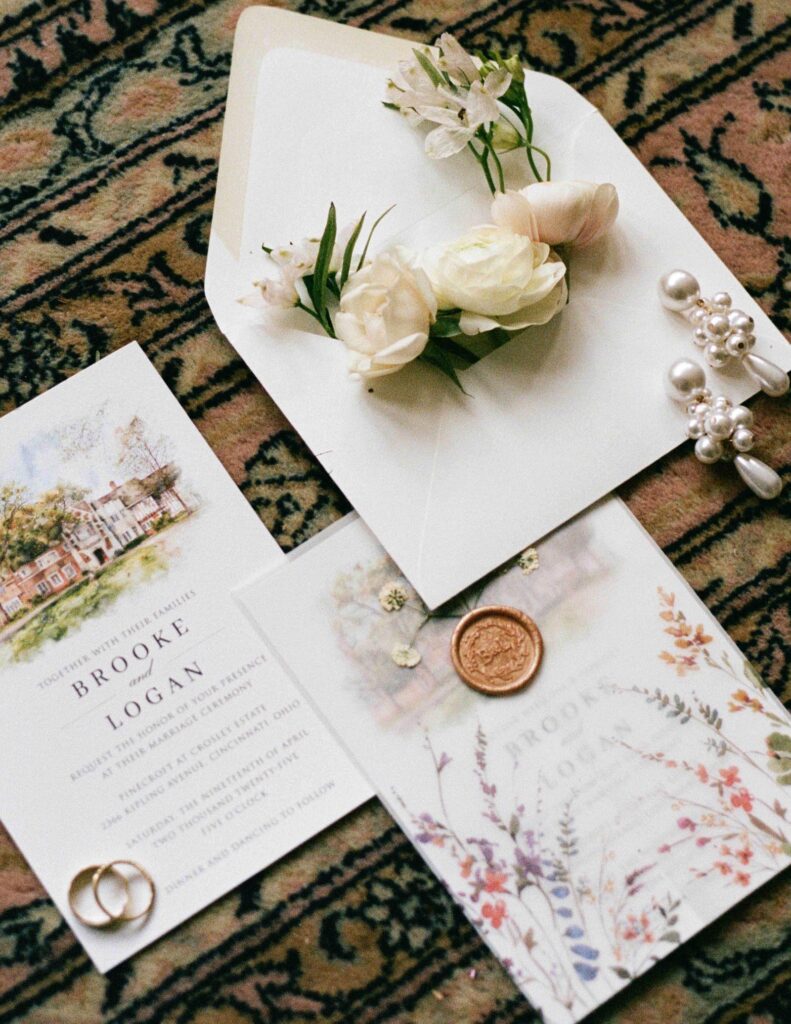 Wedding invitations on a patterned rug featuring elegant floral designs. White flowers and pearls add a soft, romantic touch.