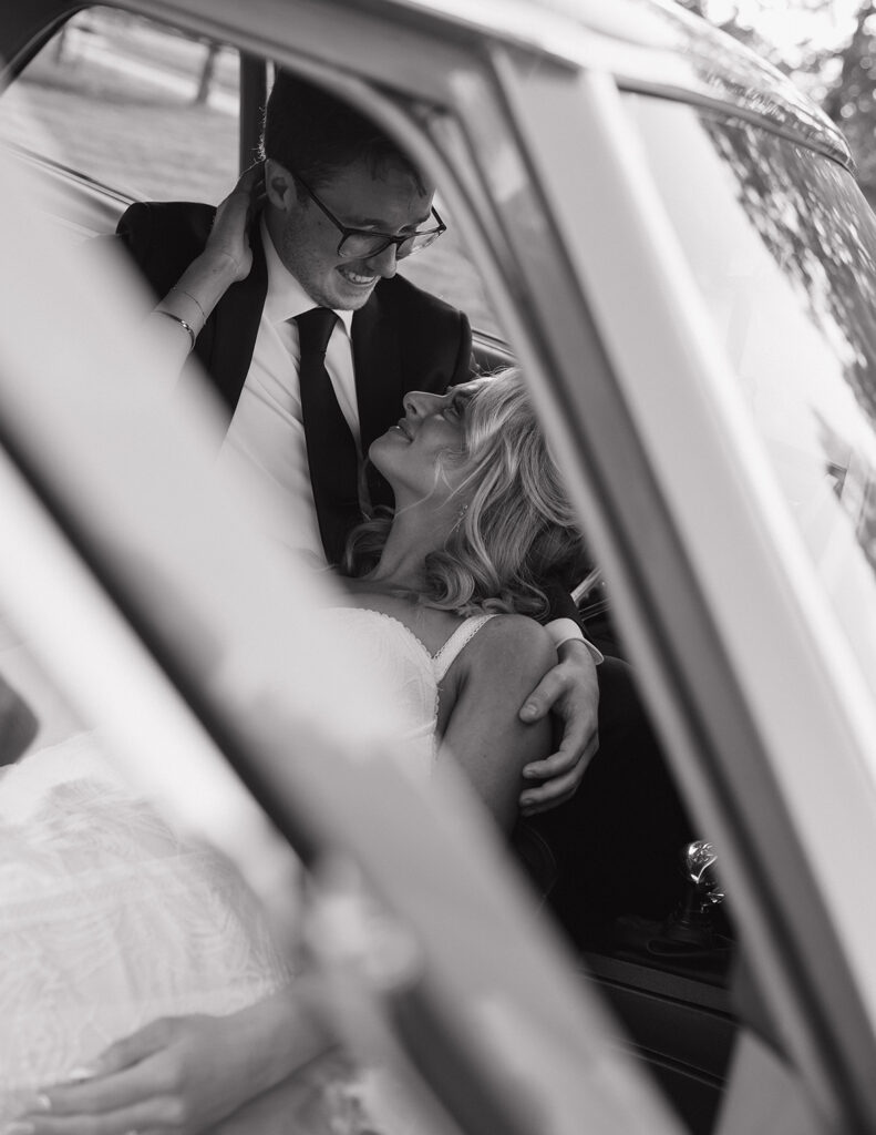 A couple sits inside a car, gazing lovingly at each other. The man in a suit holds the woman in a white dress. The mood is romantic.
