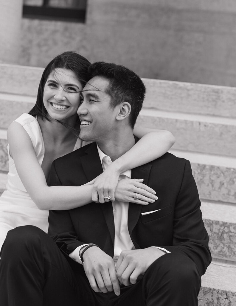 Mary and Kato sit on steps, with the woman in a white dress embracing the man in a black suit. Both are smiling, conveying love and happiness.