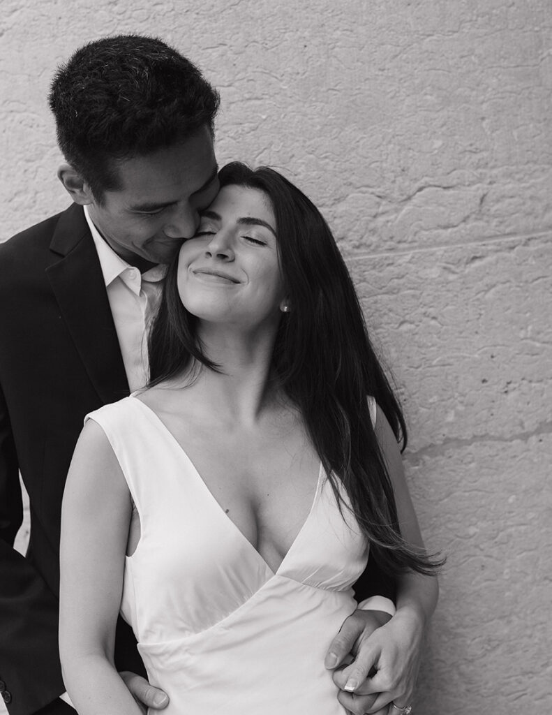 A couple in formal attire embraces, sharing an intimate moment. The man gently kisses the woman's temple. The scene conveys love and tenderness against a textured wall.
