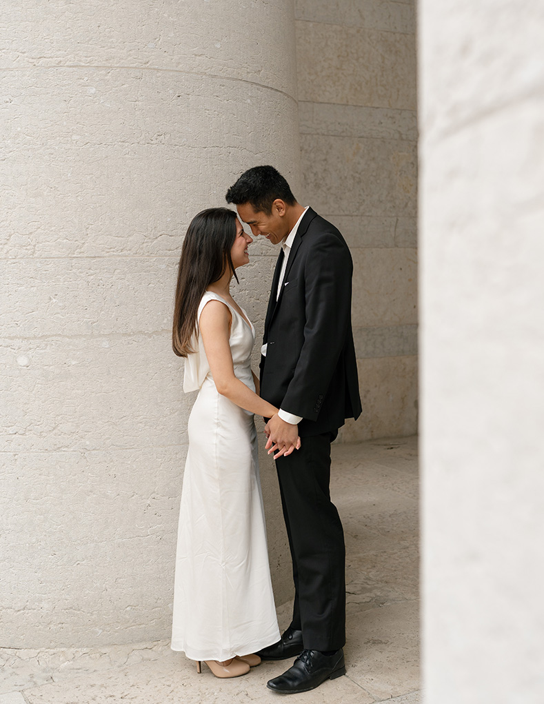 Columbus, Ohio couple stands closely, holding hands in front of a large stone pillar. The woman wears a white dress; the man is in a black suit. They look lovingly at each other.