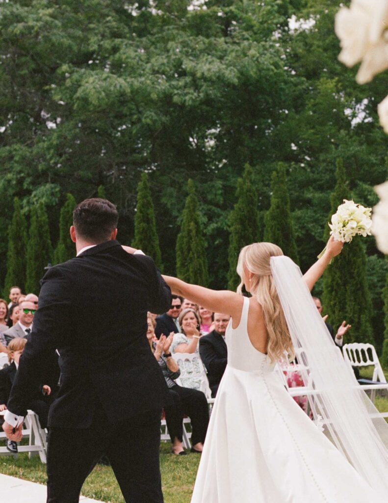 A bride and groom happily walk down the aisle outdoors. The bride holds a bouquet aloft, surrounded by clapping guests and lush greenery in the background.
