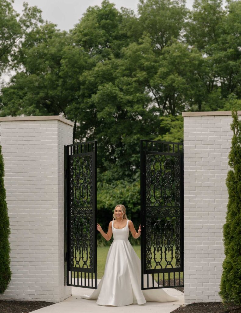 A woman in a white wedding dress stands between open black wrought-iron gates framed by white brick walls, lush green trees in the background.
