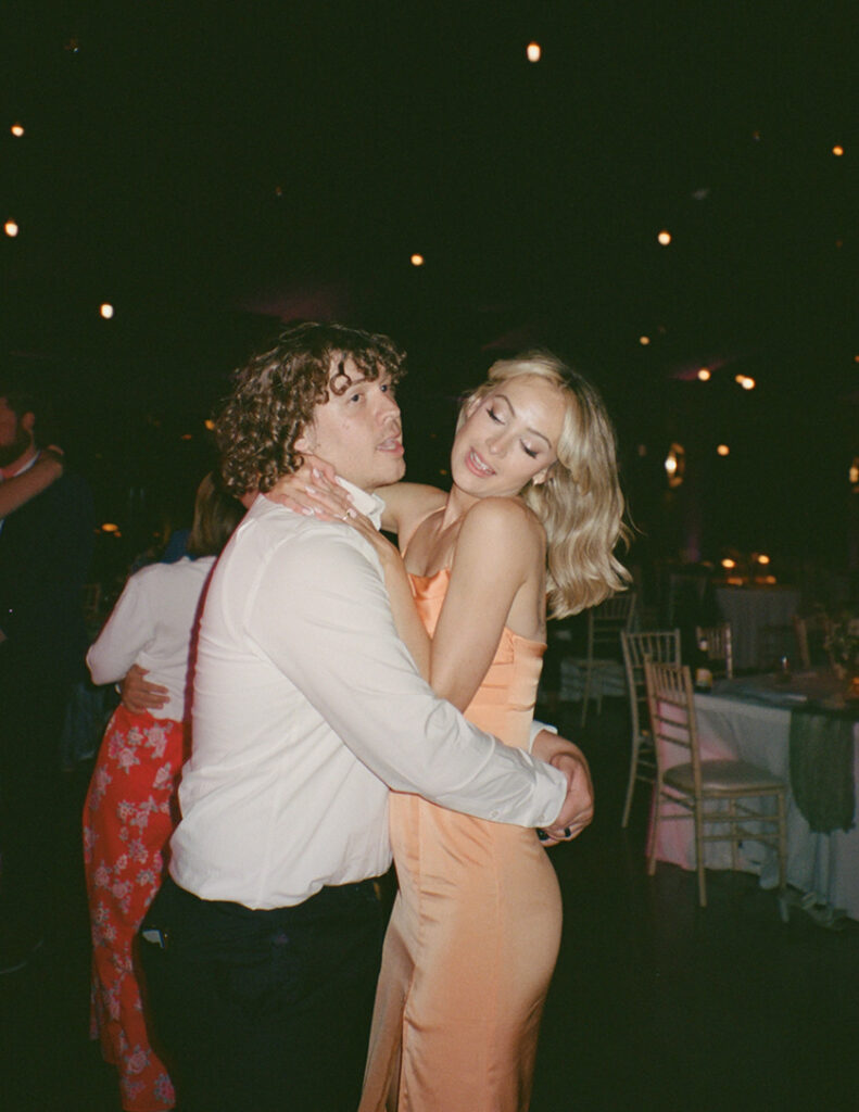 A couple in a joyous dance embrace at a lively event. The woman wears an elegant peach dress, and the man is in a white shirt, both smiling amid soft ambient lighting.