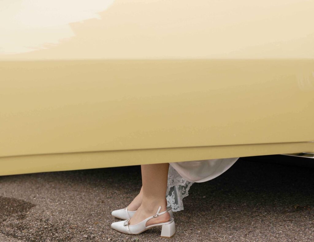 Partial view of a pale yellow vintage car with a woman stepping out, showing white block-heeled shoes and a hint of a lace dress, suggesting elegance.