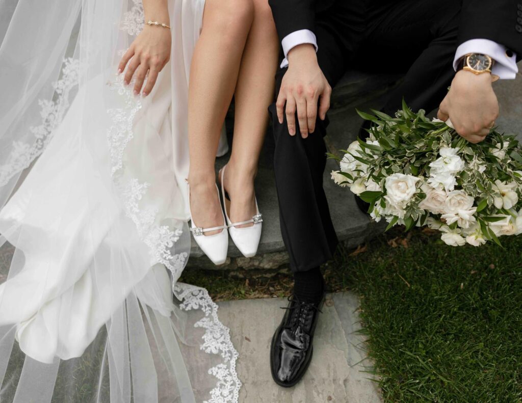Bride in white heels and lace dress sits beside groom in black suit, holding bouquet of white flowers. Romantic, elegant wedding atmosphere.