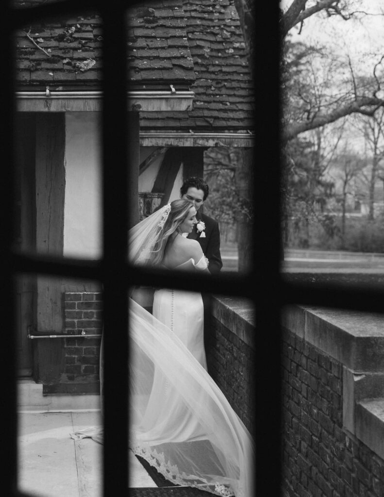 Black and white photo of a bride and groom sharing an intimate moment outside a rustic building, framed through dark window panes, conveying romance and timelessness.