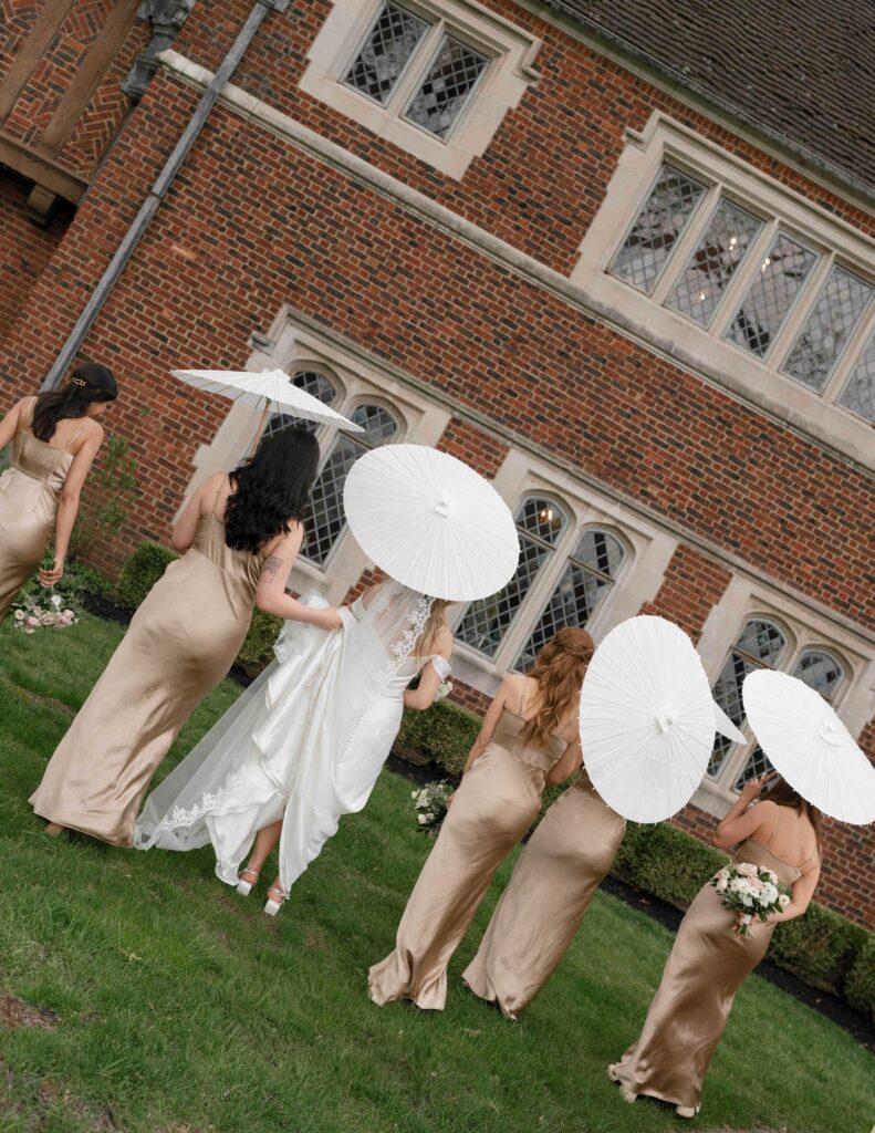 Bridesmaids in champagne dresses and a bride in white walk on grass with white parasols. They are near a brick building with large windows.
