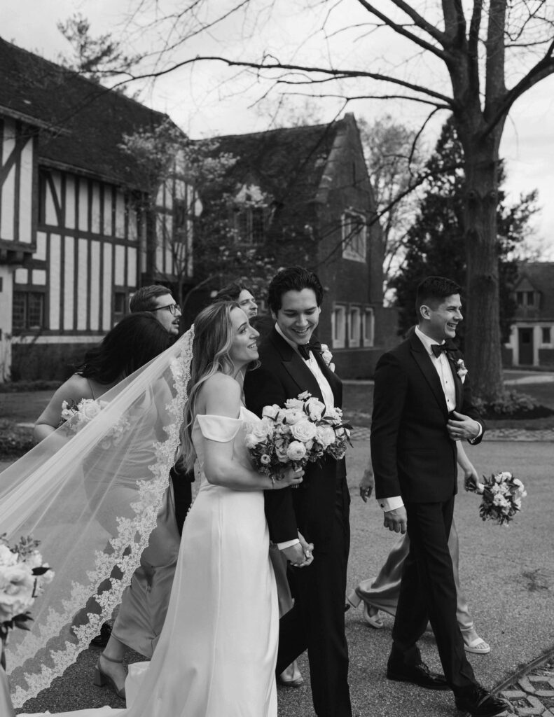 A Cincinnati, Ohio bride and groom walk outdoors with friends, the bride holding a bouquet and veil flowing. They all wear formal attire, smiling warmly.