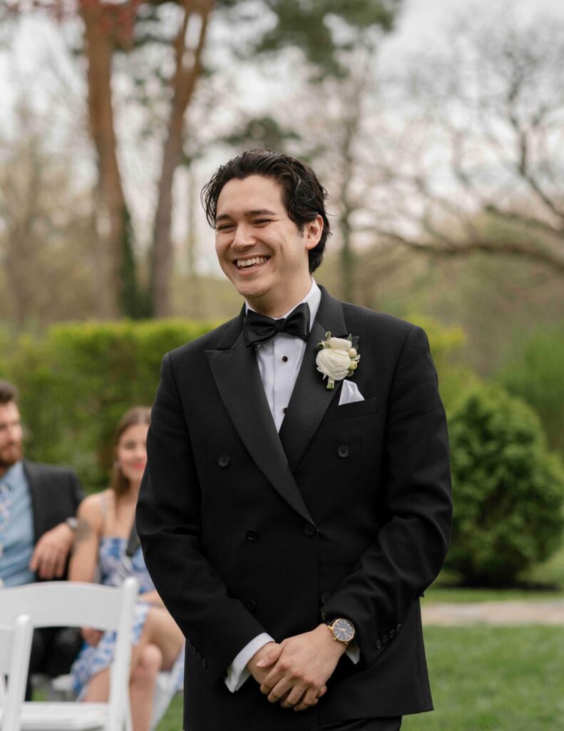 Cincinnati groom in a black tuxedo with a white boutonniere stands outdoors, surrounded by trees and guests in a garden setting.