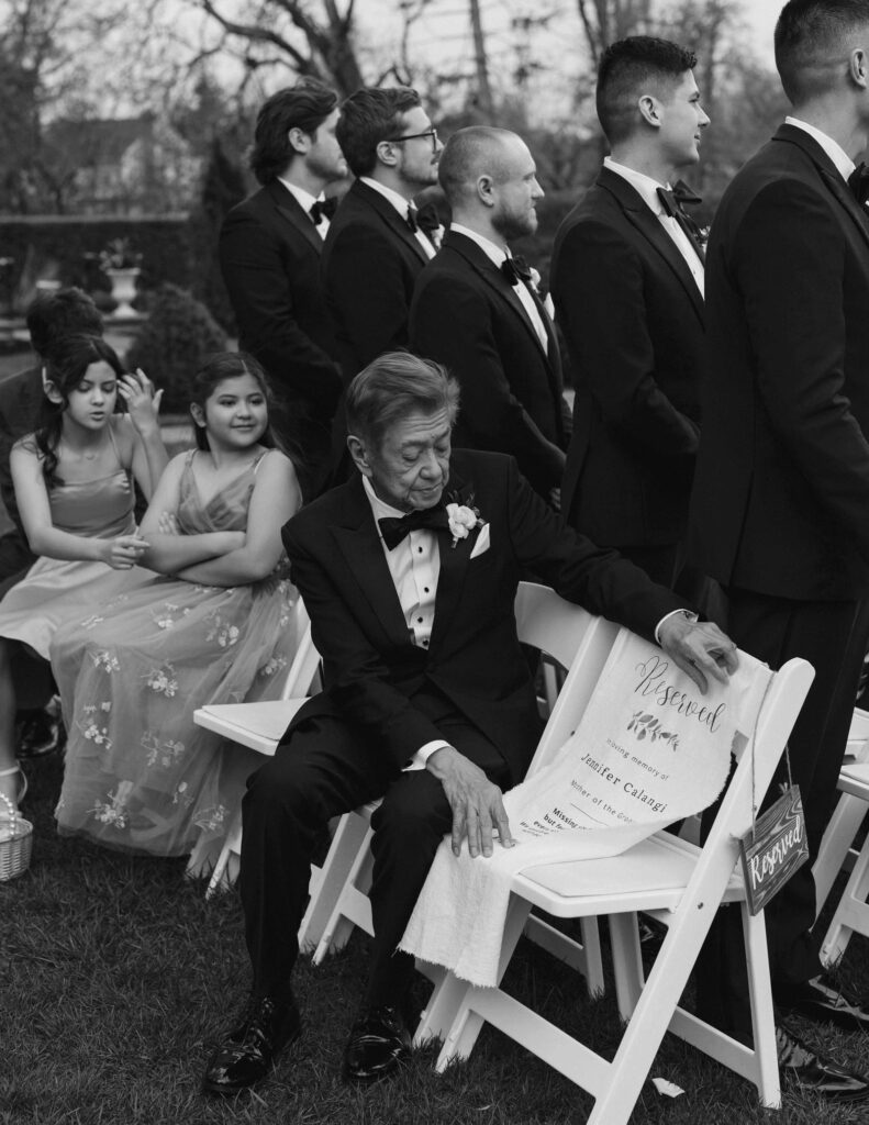 A black-and-white image of a wedding scene. An elderly man in a tuxedo sits on a reserved chair while others stand nearby. The mood is formal and reflective.