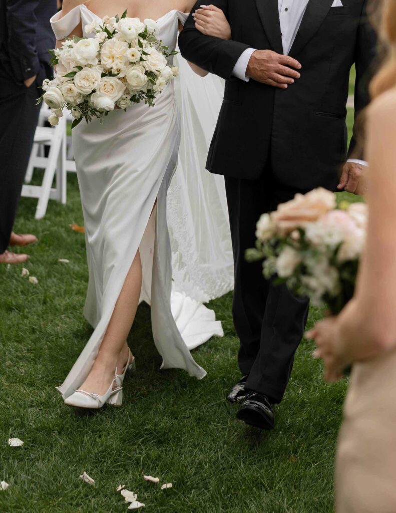 Bride in a white gown with a bouquet of white roses walks arm-in-arm with a man in a suit on grass, wedding guests holding flowers nearby.