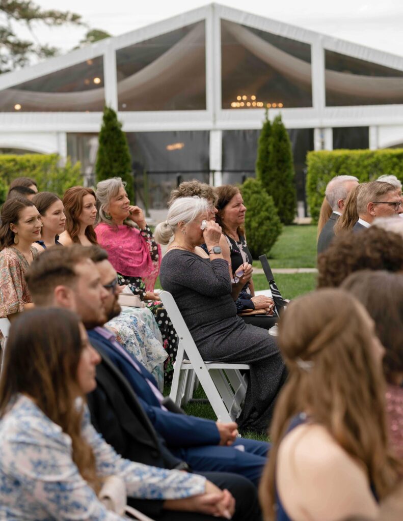 A diverse group of wedding guests sit outdoors on white chairs, some visibly emotional. A large, elegant white tent is in the background.
