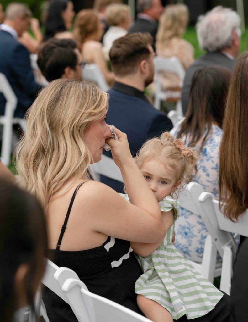 A woman in a black dress sits among a crowd at an outdoor event, wiping tears with a tissue. A young child in a striped dress sits on her lap.