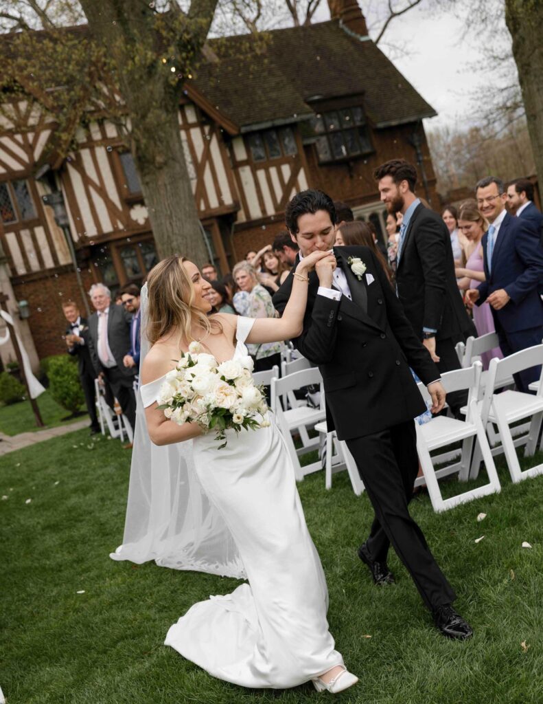A Cincinnati bride in a white dress holds a bouquet while walking arm-in-arm with the groom, who kisses her hand. Guests cheer behind them outside a Tudor-style house.