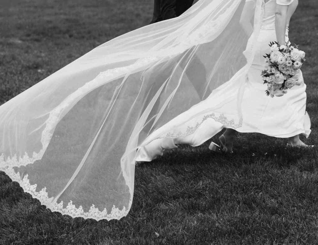 Black and white image of a bride walking on grass, her long veil flowing elegantly. She holds a bouquet of roses, conveying a sense of grace and joy.