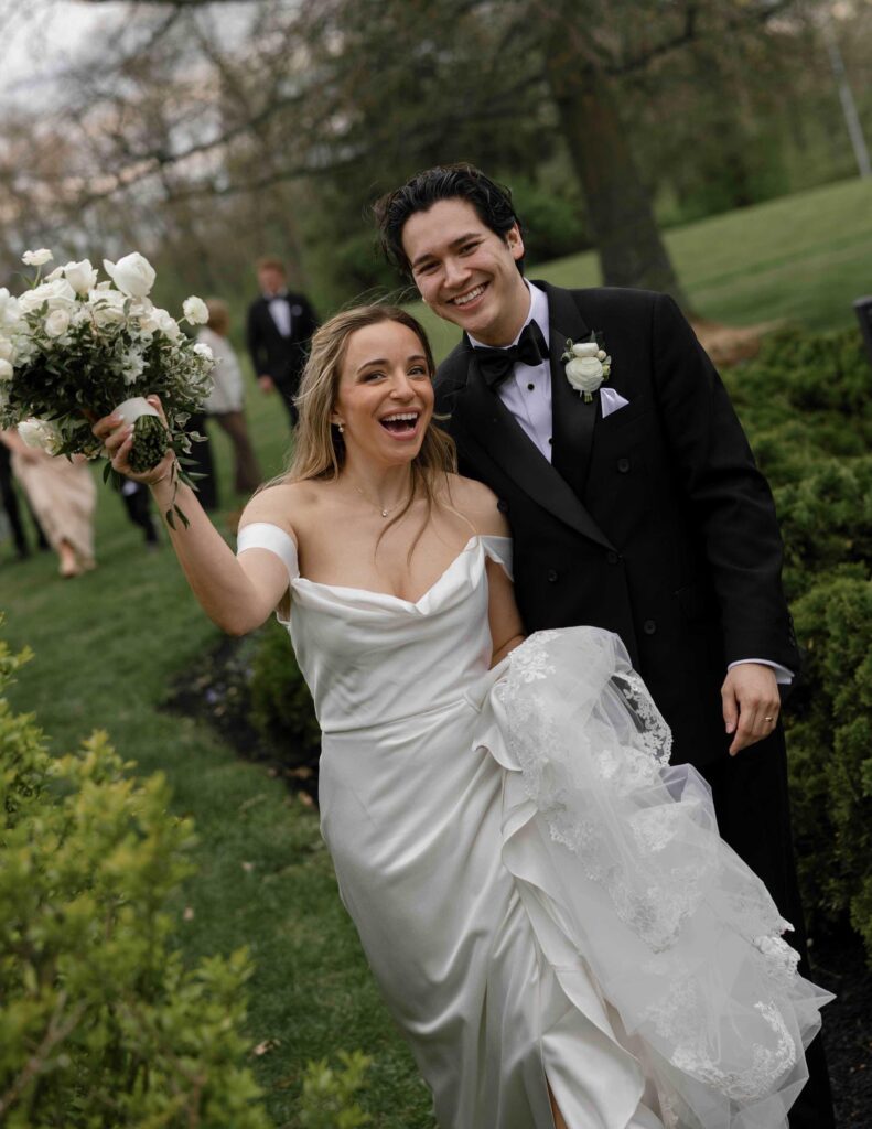 A happy bride and groom in a garden setting. The bride, in a white gown, holds a bouquet of white flowers, while the groom wears a black tuxedo, both smiling widely.