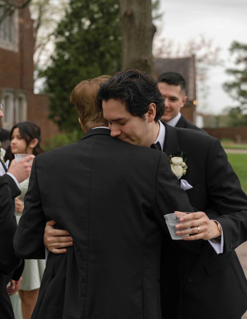 Two men in formal black suits embrace at an outdoor event. One holds a plastic cup, and a boutonniere is visible. The atmosphere is warm and emotional.