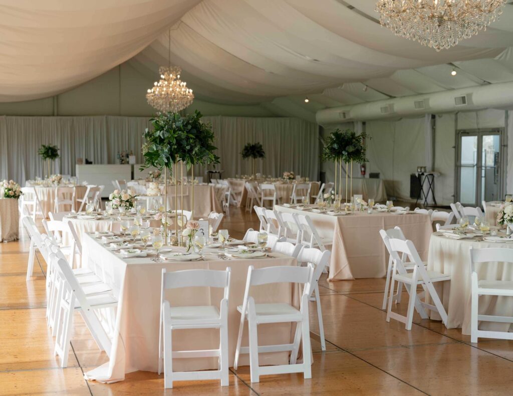 Elegant wedding reception at Pinecroft Mansion in Cincinnati with white decor and chandeliers. Tables are set with white chairs, cream linens, and tall green centerpieces, creating a sophisticated atmosphere.