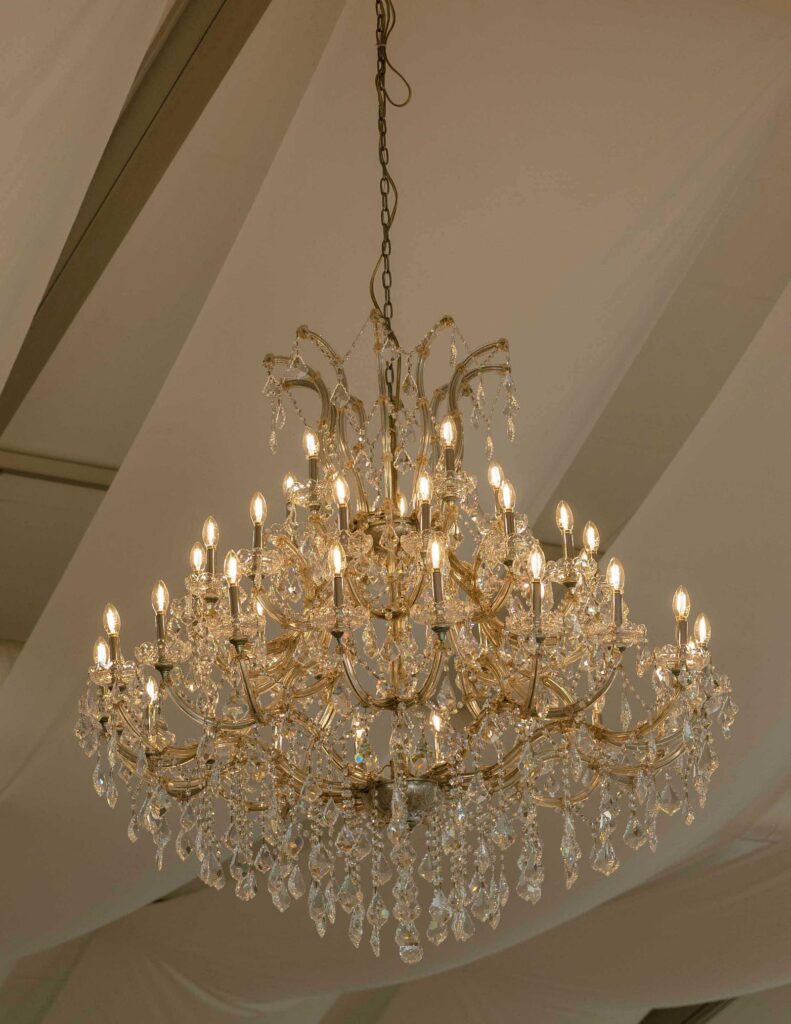 Elegant crystal chandelier with glowing lamps hangs from a ceiling, casting a warm, luxurious glow. The atmosphere is opulent and refined.