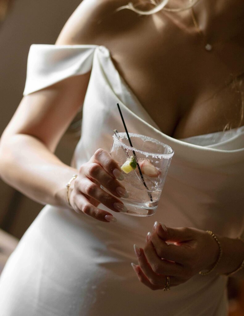 A bride in an elegant white dress holds a cocktail glass garnished with a lime. The mood is sophisticated, with soft lighting accentuating her attire.