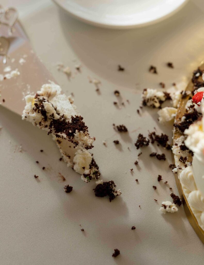 Wedding cake crumbs and frosting scattered on a tablecloth with a cake server and a slice missing from the plate, conveying a sense of messiness and indulgence.