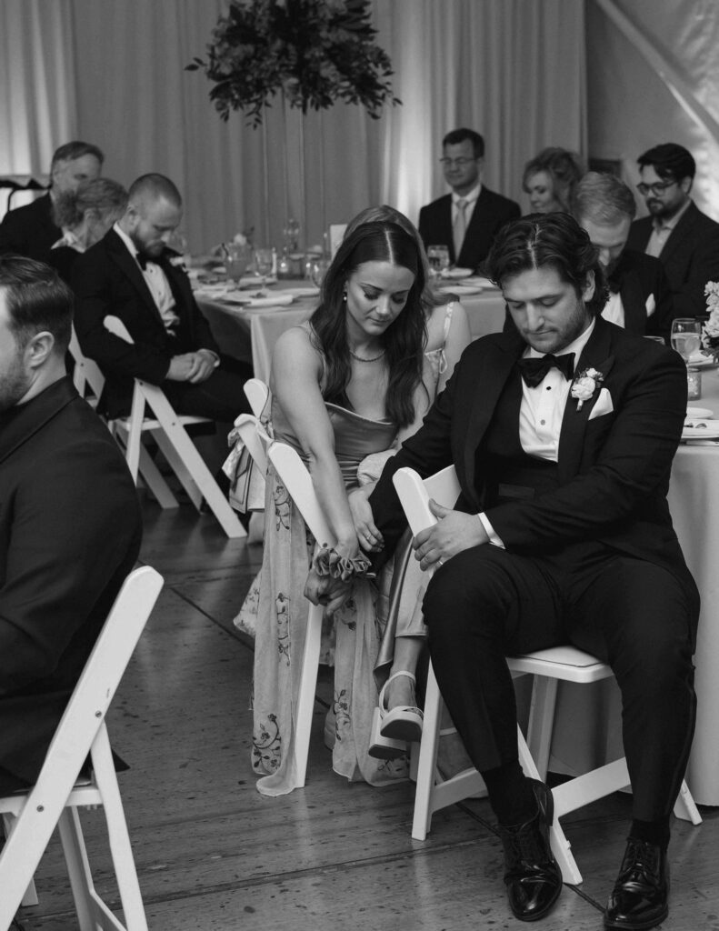 A monochrome image of a formal event. A couple sits closely on white chairs, holding hands, appearing thoughtful. Attendees sit around tables with a calm ambiance.