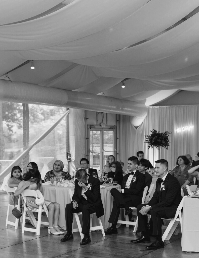 Guests seated at a wedding reception under draped fabric ceiling. People in formal attire appear reflective or emotional, setting a heartfelt tone.