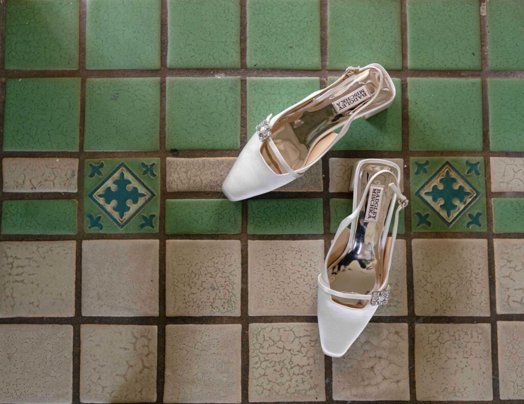 Elegant white high-heel shoes with jeweled embellishments lie on a vintage green and beige tiled floor, conveying a sense of sophistication.