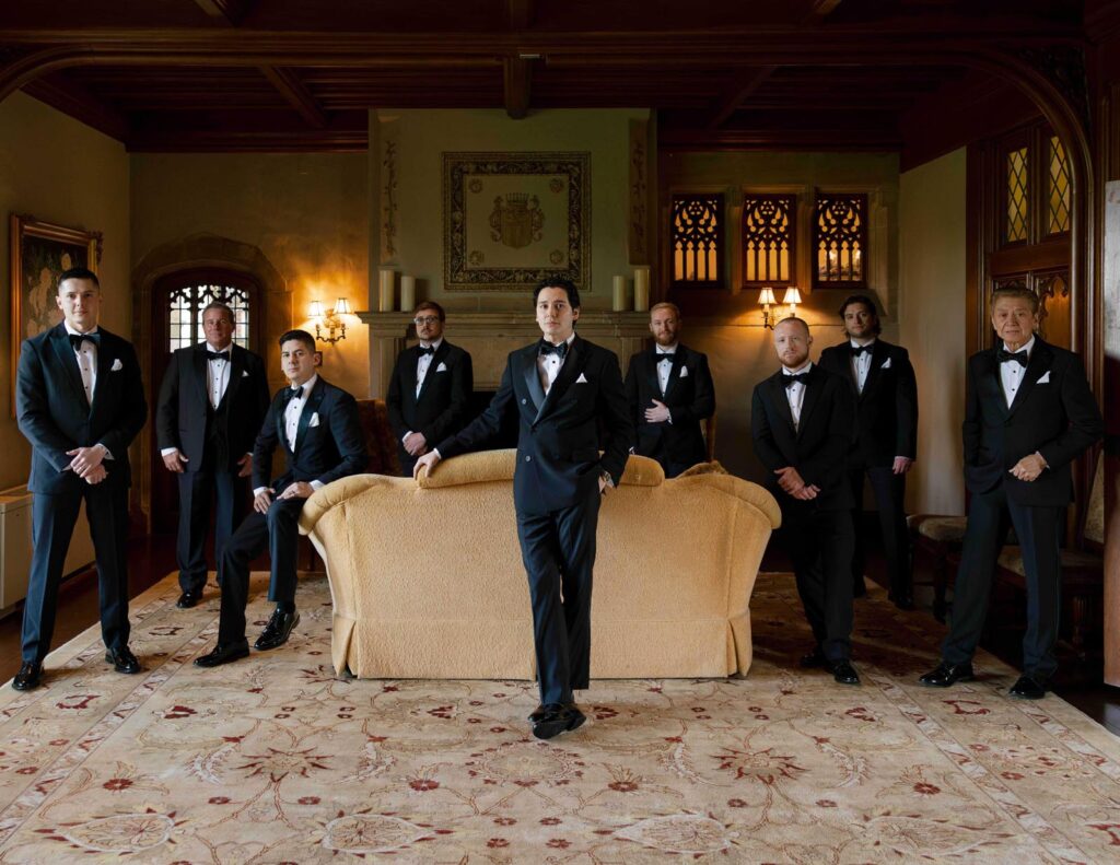 A group of eight men in tuxedos stand confidently in an elegant, dimly lit room with wood paneling, a beige sofa, and an ornate carpet. Mood is formal.