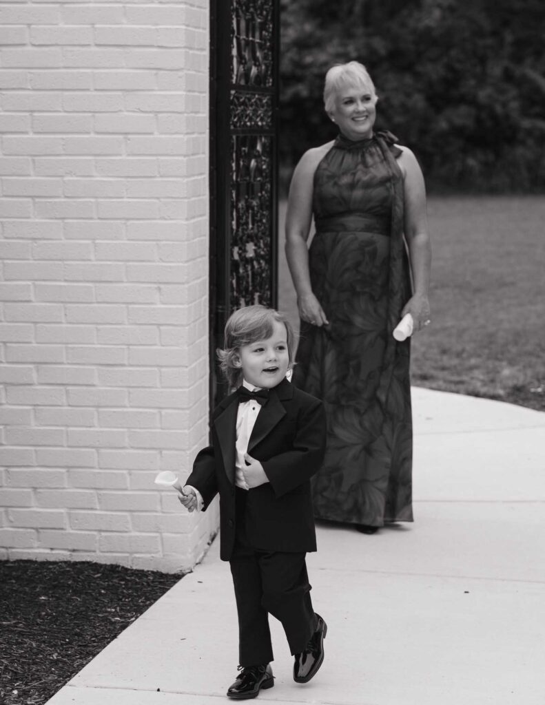 A young boy in a tuxedo walks on a path holding a rose petal, smiling. Behind him, a woman in a patterned gown also holds a petal, smiling warmly.