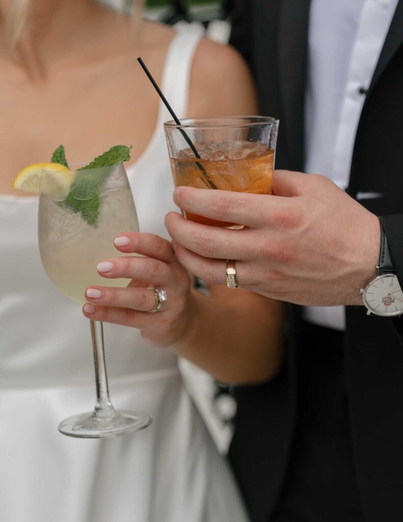 A couple in formal attire holding drinks; the woman has a lemon and mint cocktail, and the man has a whiskey with ice. Both display wedding rings.