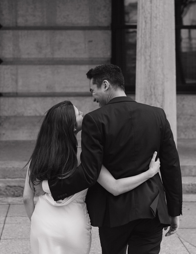 A couple in formal attire, a man in a suit and a woman in a dress, walk closely arm-in-arm up a stone staircase. They smile warmly at each other.