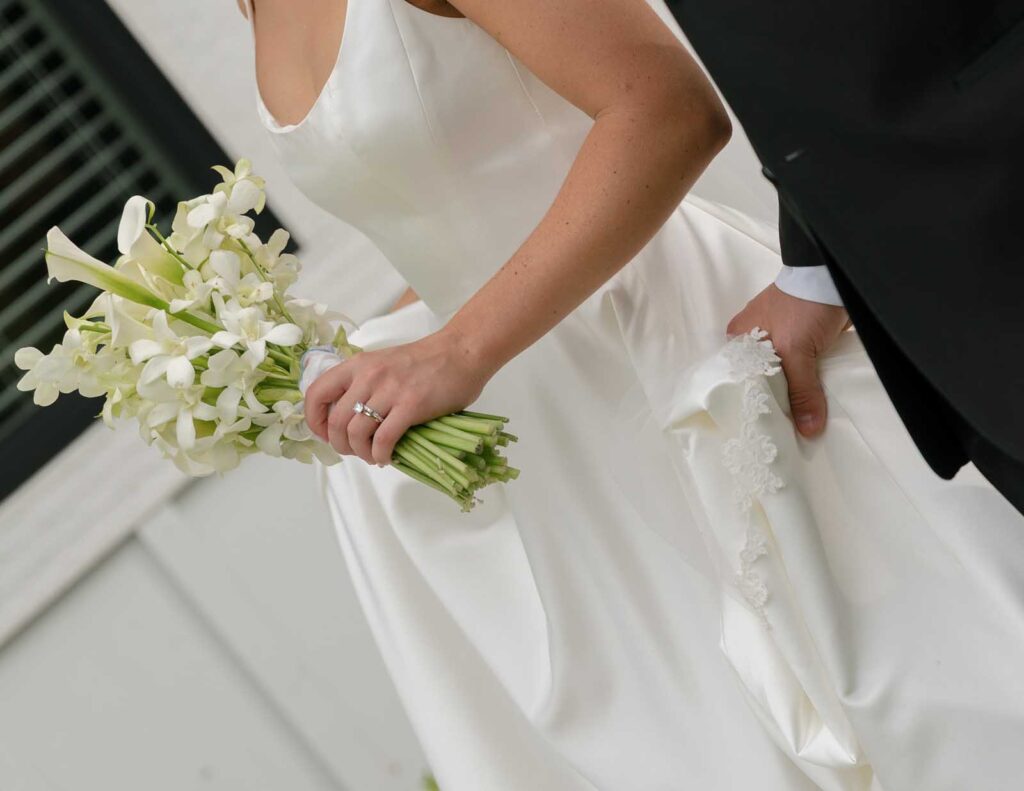 Bride in a white dress holds a bouquet of white flowers, displaying an engagement ring. A groom, partially visible, supports her dress. Elegant and romantic scene.