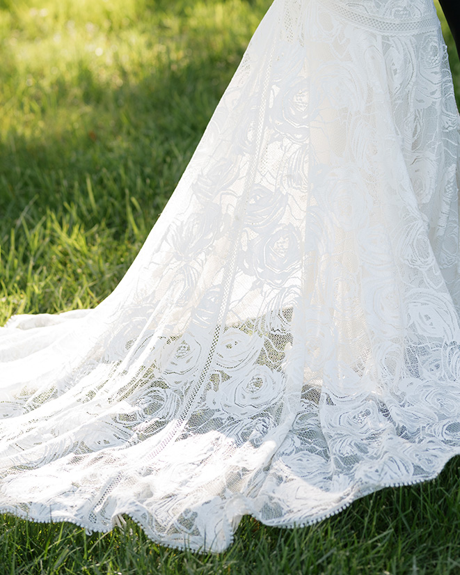 A close-up of a white lace wedding dress with a floral pattern, trailing elegantly on green grass. The scene conveys a serene, romantic feel.