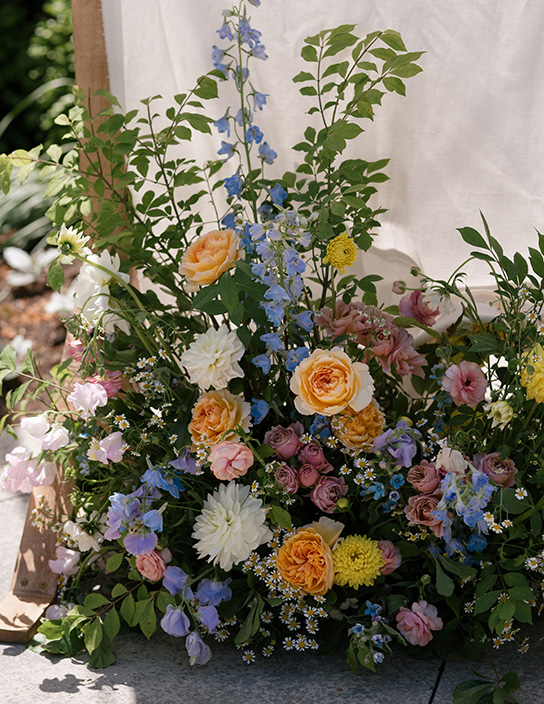 A lush arrangement of flowers with yellow, peach, pink, and white blooms, accented by blue petals and green foliage, creates a vibrant, natural display against a soft, light backdrop.