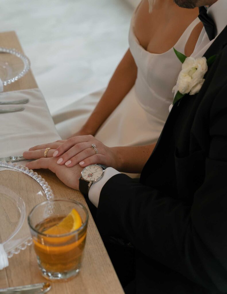 A bride and groom hold hands at a table, her white dress contrasting his black suit