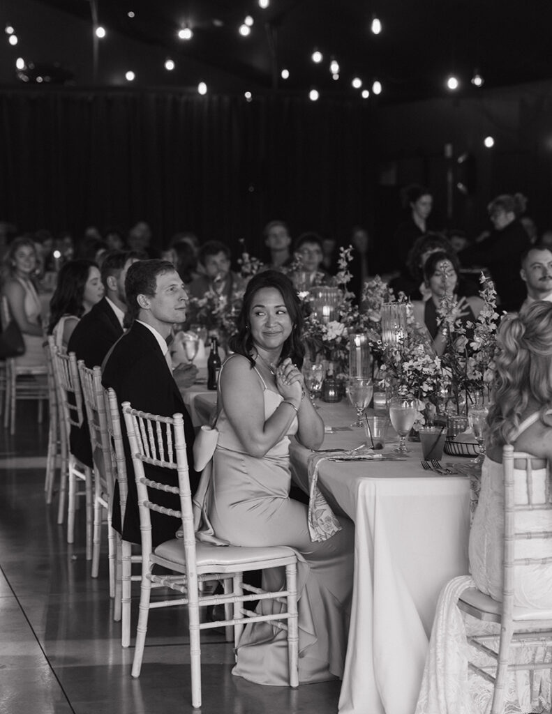 A black-and-white image of a joyous wedding reception. Elegantly dressed guests sit at decorated tables, engaged and smiling under a canopy of lights.
