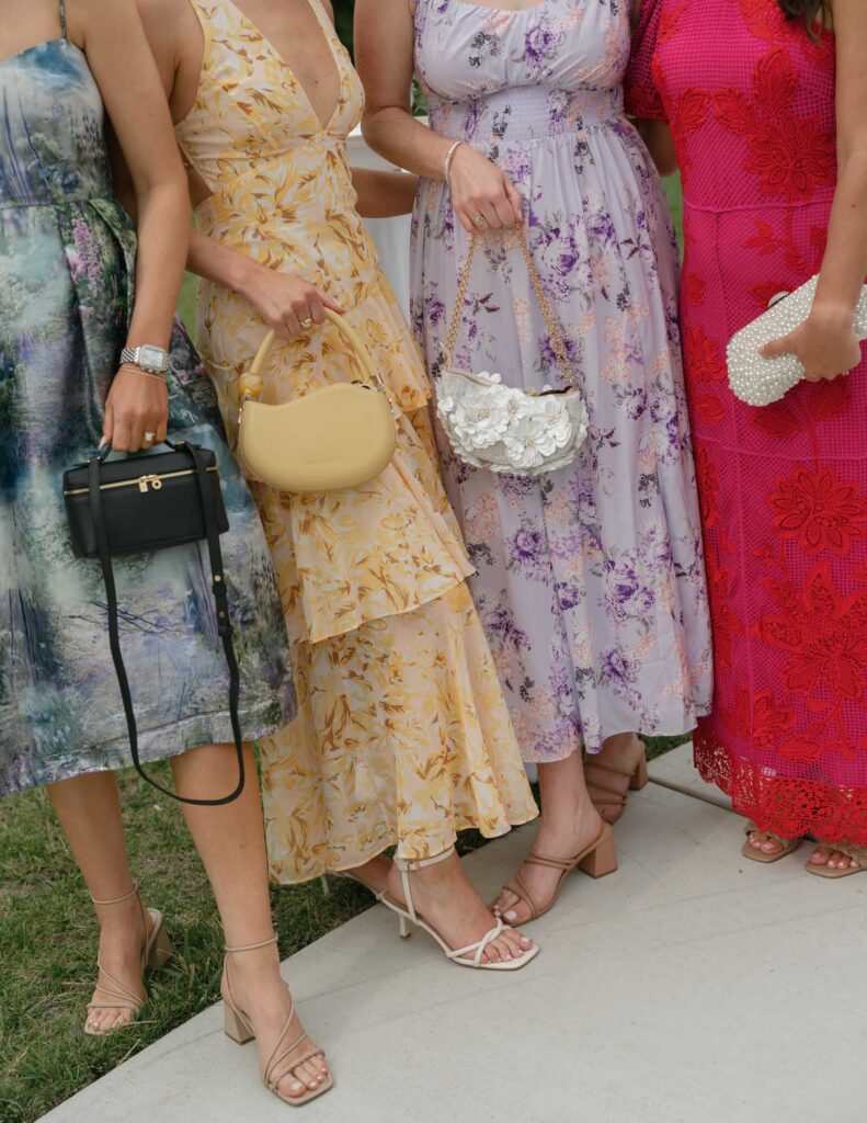 Four women in floral and lace dresses stand in a row, holding elegant handbags. Their attire features yellow, pastel purple, and red tones, evoking a summery and celebratory mood.