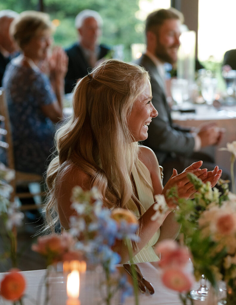 A woman with long blonde hair sits at a dinner event, smiling and clapping. The atmosphere is warm, with blurred floral decorations and people in the background.