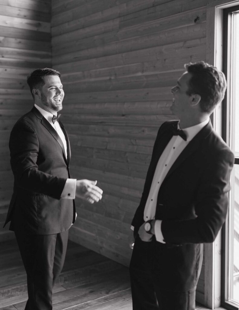 Two men in tuxedos share a joyful moment, shaking hands and smiling, in a room with wooden walls and a large window letting in natural light.
