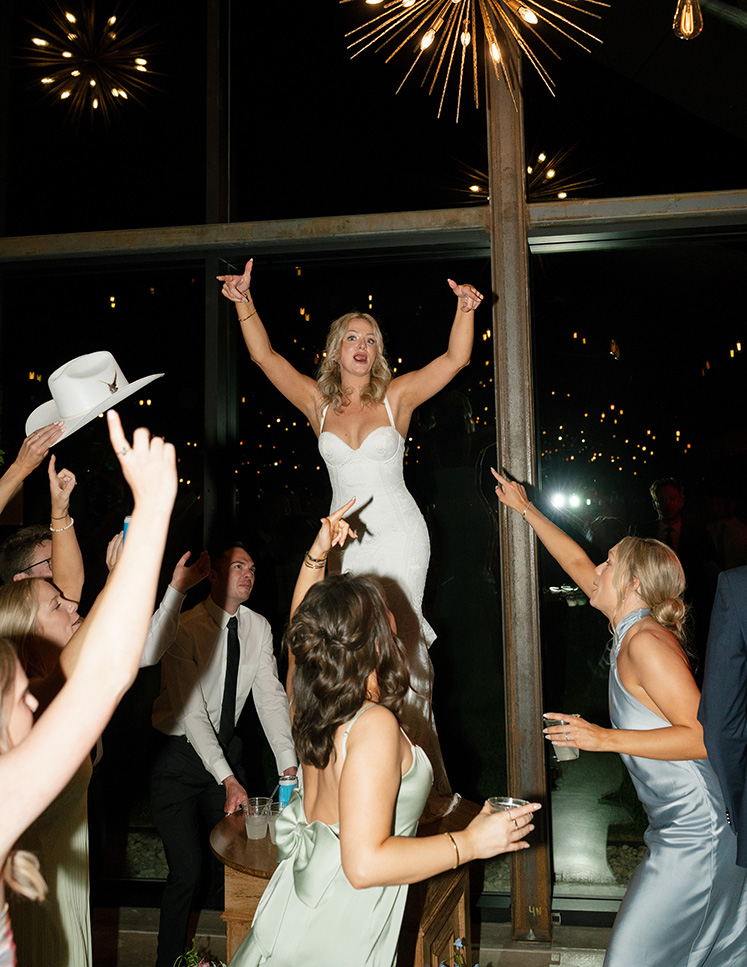 A joyful bride in a white dress stands on a table, arms raised, surrounded by cheering guests at a lively wedding reception with a modern chandelier.